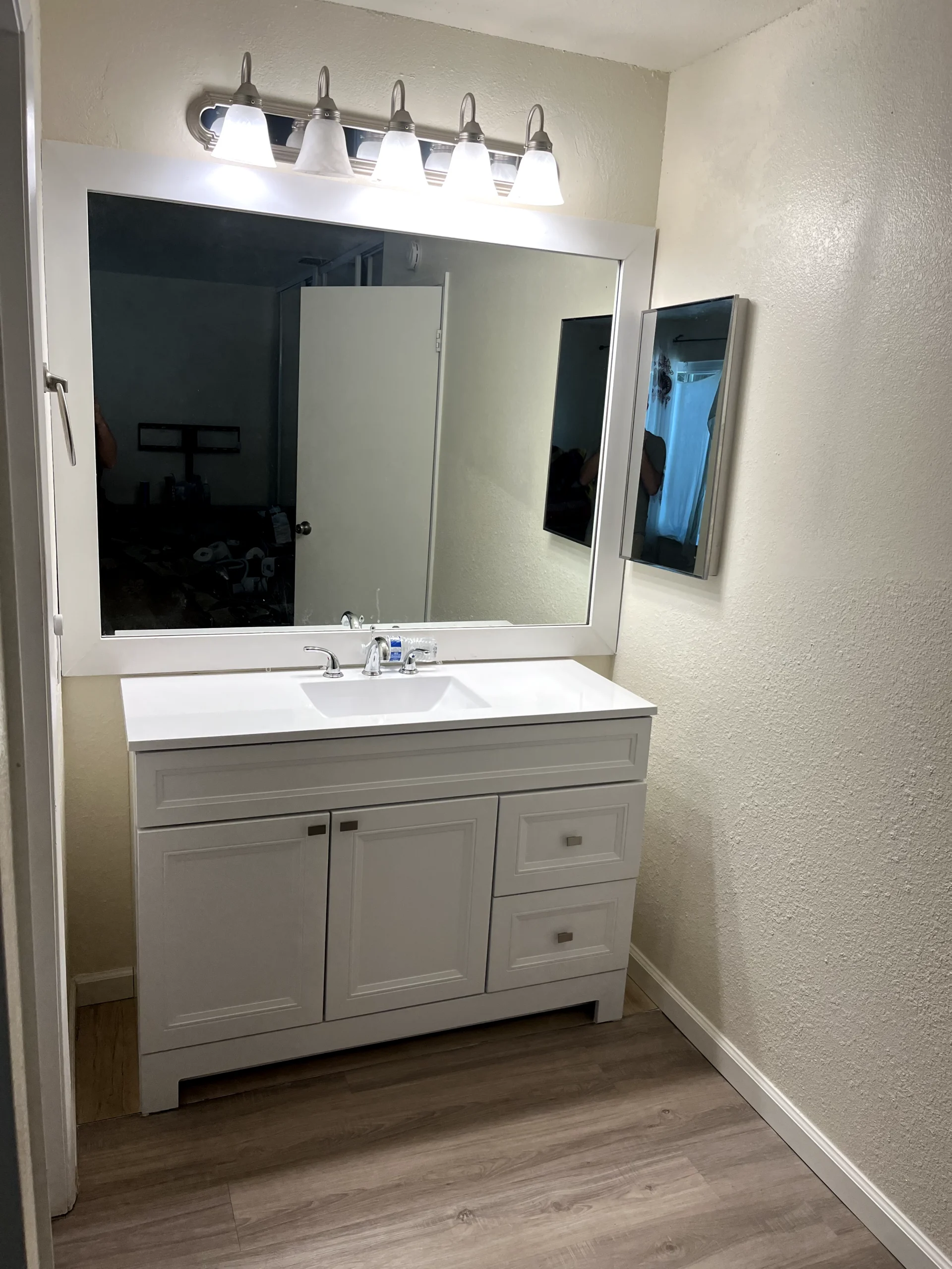 Bathroom vanity remodel in Sacramento with white cabinet, updated sink, mirror, lighting, and wood-look flooring.