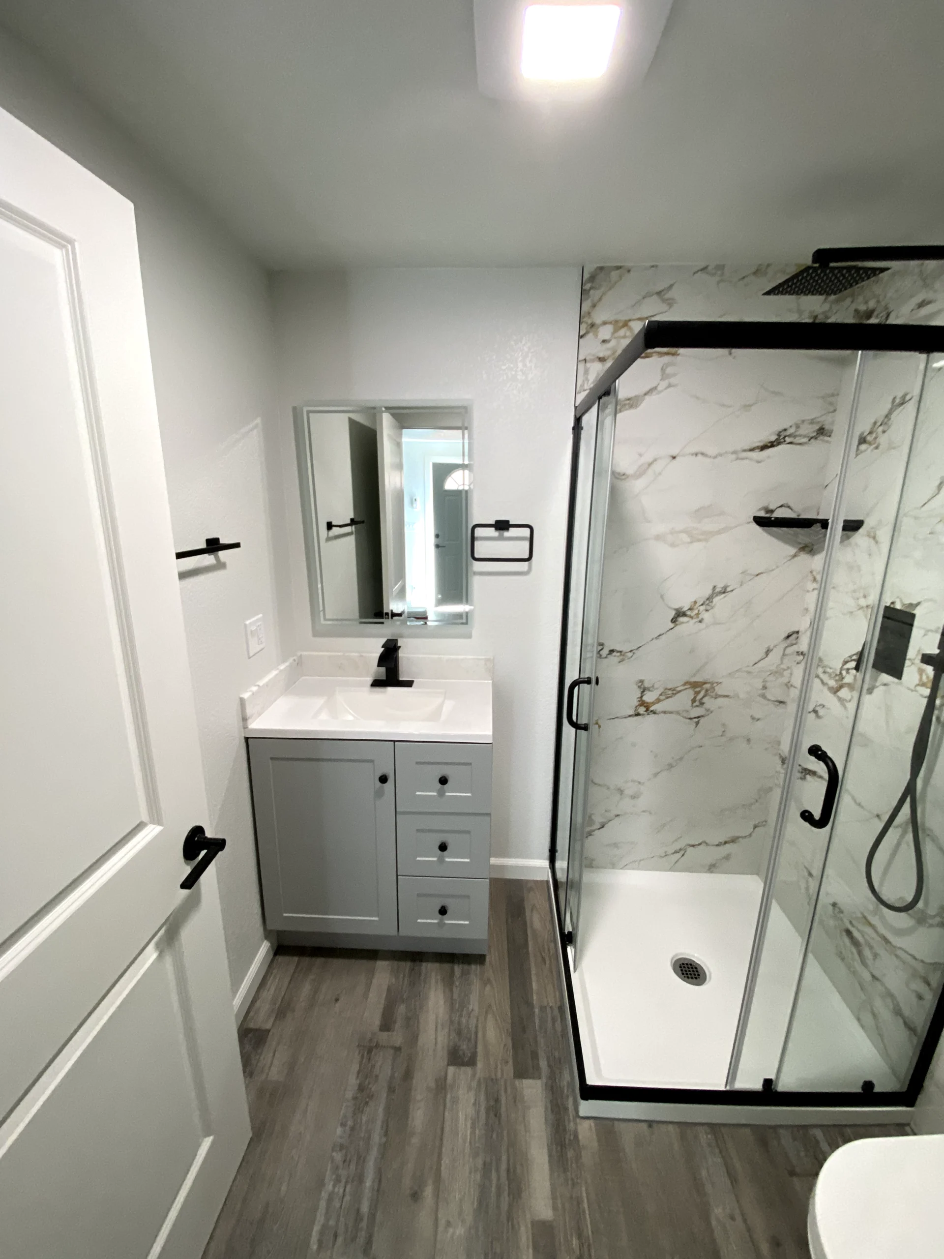 Compact bathroom remodel in Orangevale with glass shower doors, marble-look shower walls, gray vanity, and wood-look flooring.