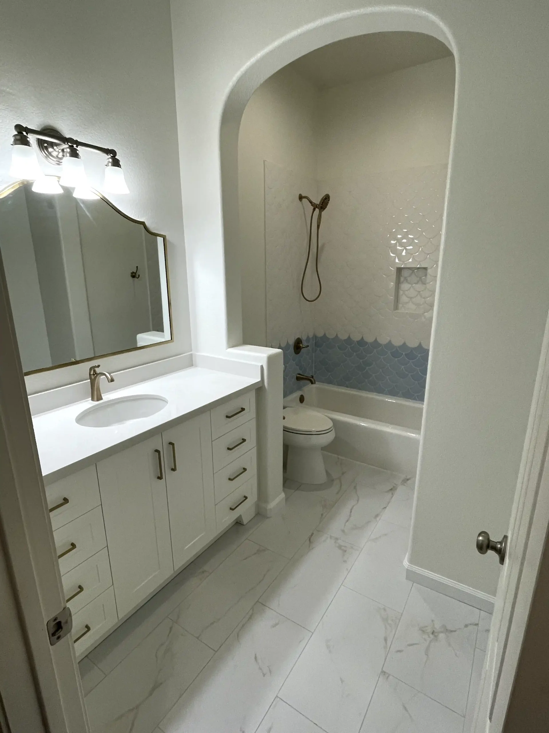 Bright guest bathroom remodel in Fair Oaks with white vanity, brass hardware, marble-look flooring, arched tub area, and blue accent tile.