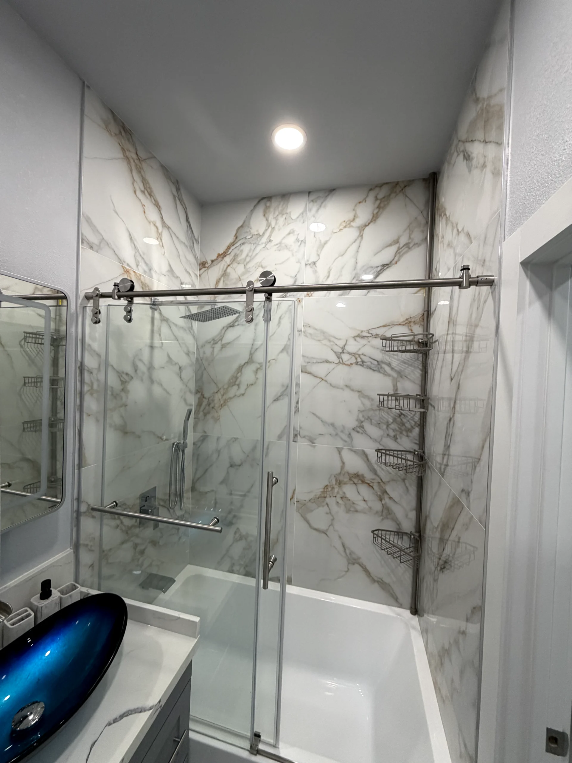 Walk-in shower remodel in Elk Grove with marble-look wall tile, sliding glass door, rainfall shower head, and built-in storage.